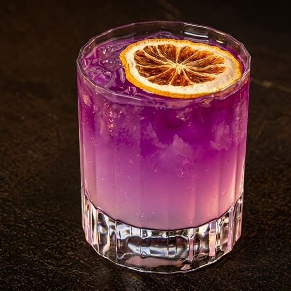 A vibrant purple cocktail with ice and a dried orange slice garnish, on a dark textured surface.